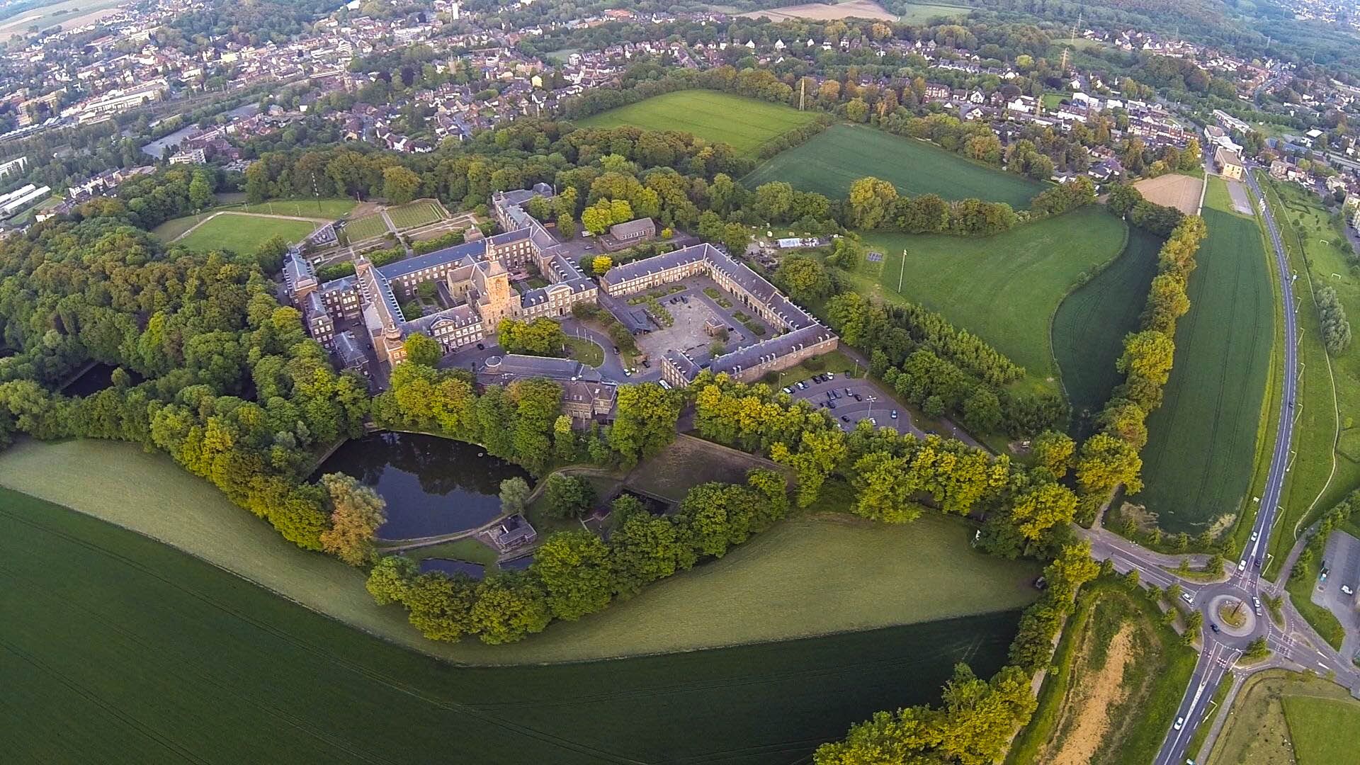 An aerial view of a large estate with a central courtyard, surrounded by fields and a lake.