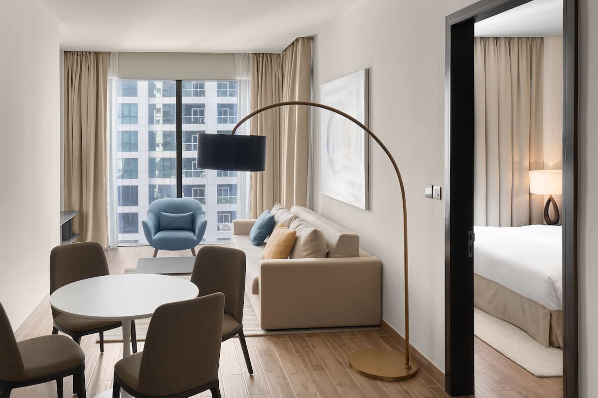 Een moderne hotelkamer met een groot raam, een ronde eettafel, een bank, een blauwe fauteuil en een bed.