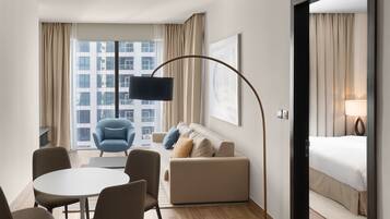 A modern hotel room with a large window, a round dining table, a sofa, a blue armchair, and a bed.