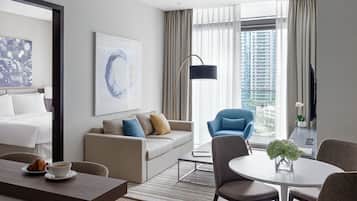 A modern hotel room with a bed, a sofa, a dining table, and a view of the city through large windows.