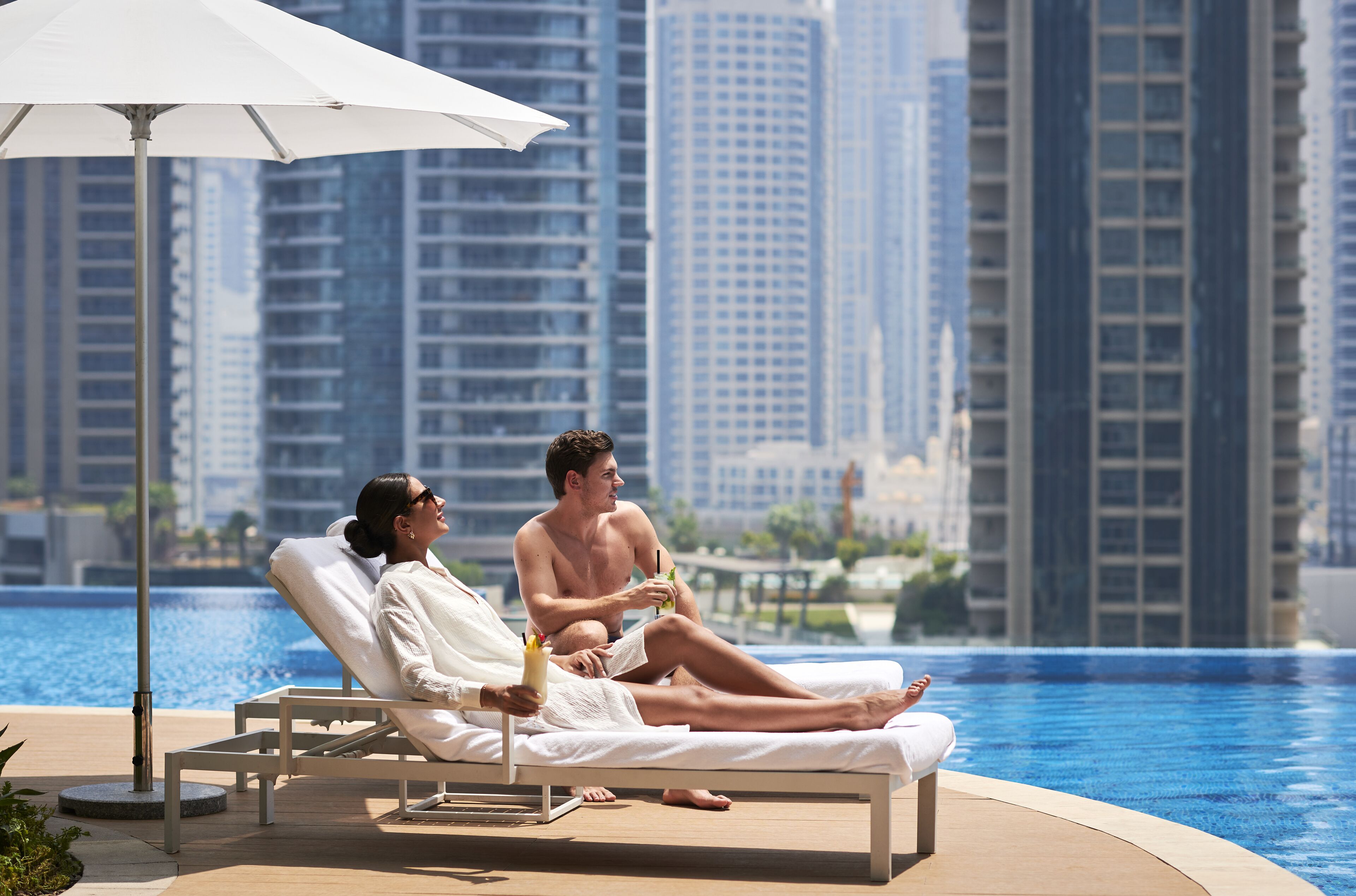 Un couple se détend près d’une chaise longue au bord d’une piscine, avec une vue sur la ville en arrière-plan.