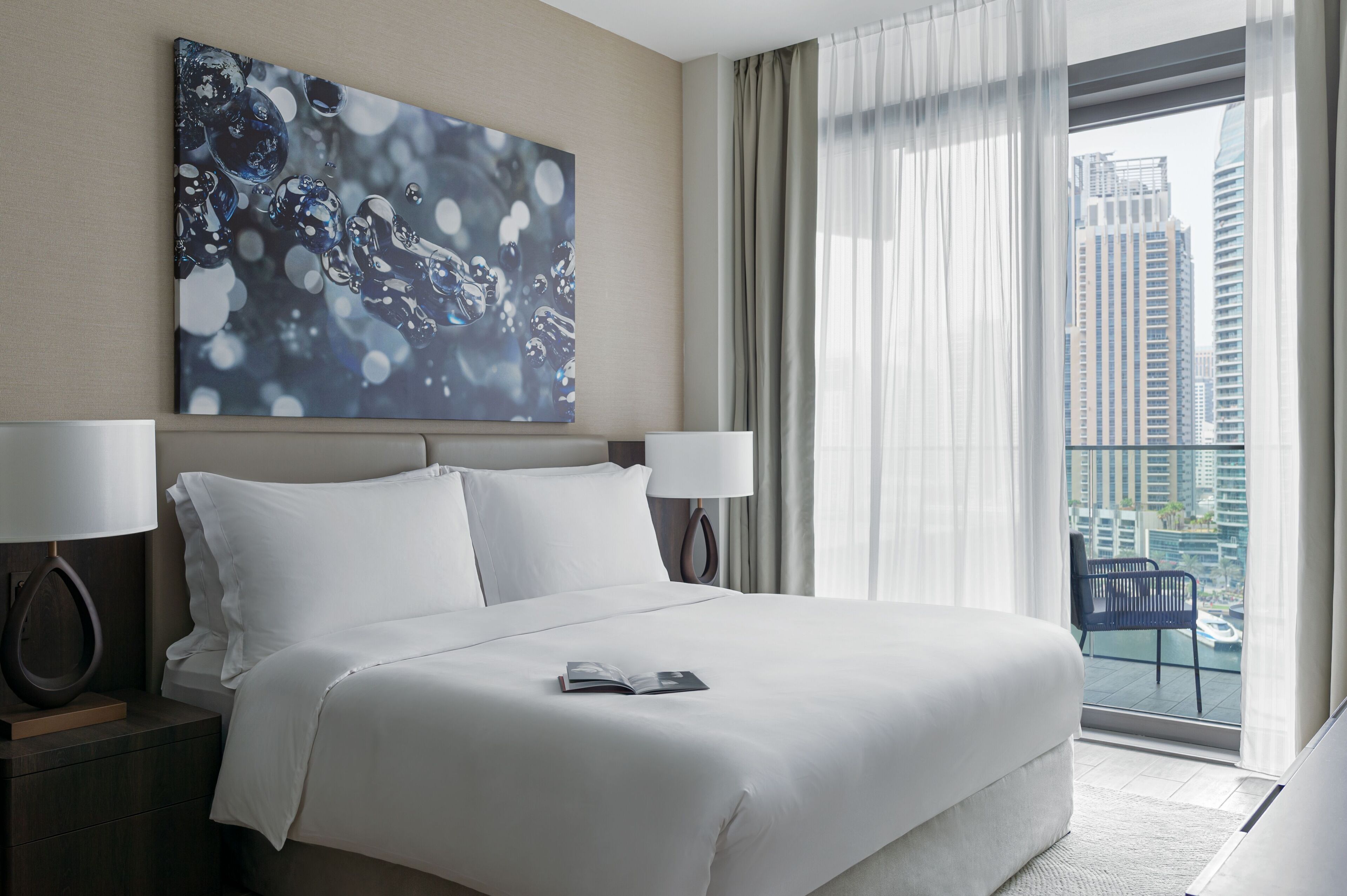 Une chambre d’hôtel moderne dotée d’un grand lit, de deux lampes de chevet, d’un balcon offrant une vue sur la ville et d’une peinture abstraite contemporaine accrochée au mur.