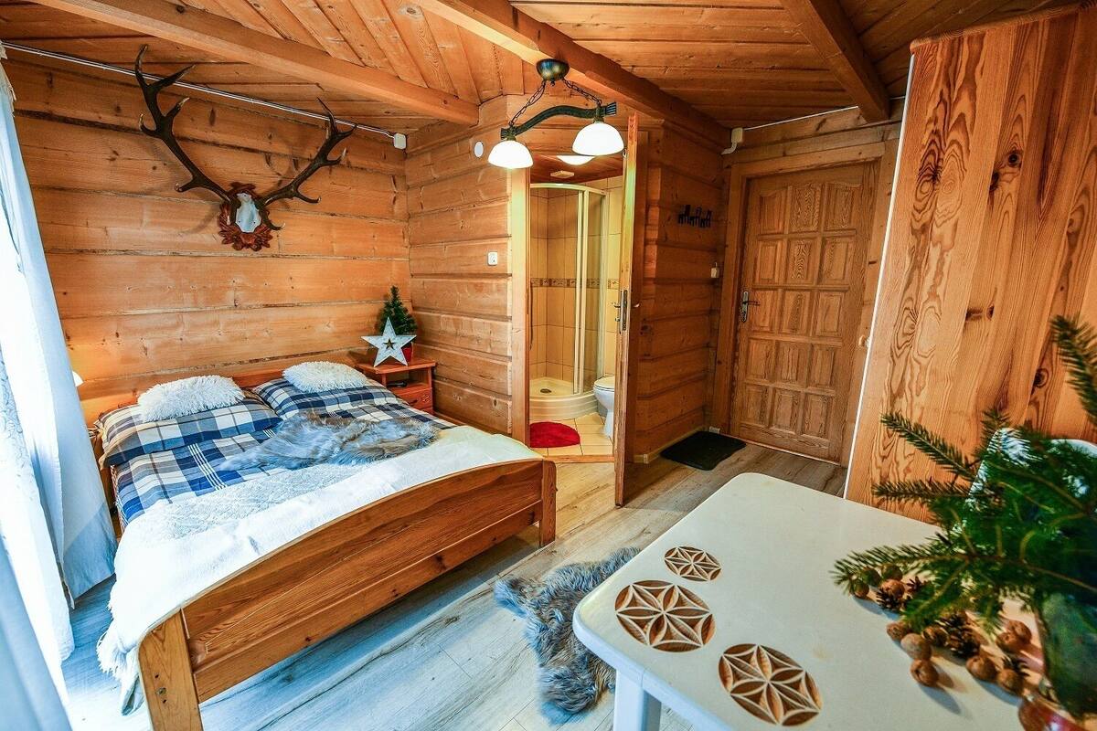 A wooden cabin interior with a bed, a small table, and a bathroom visible through an open door.