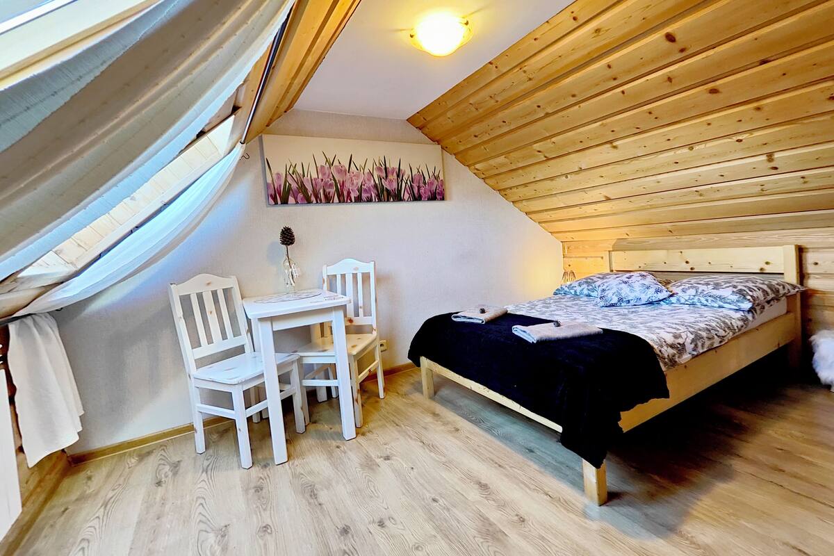 A cozy attic bedroom with a wooden ceiling, a bed, a small table with chairs, and a wall-mounted painting.