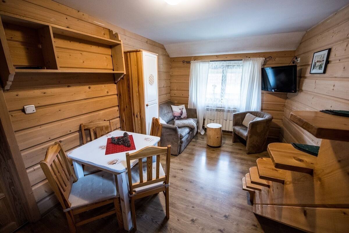 A cozy cabin interior with wooden walls, a dining table with chairs, a sofa, a round coffee table, and a television mounted on the wall.