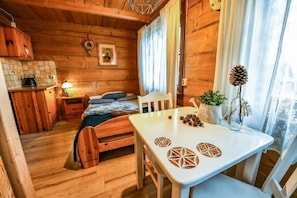 A cozy cabin interior with a wooden bed, a dining table with geometric coasters, a small kitchen area, and a window with curtains. - Willa Bachledowka (Zakopane)