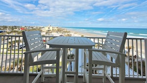 A balcony with two white chairs and a small table, overlooking a beach with a clear sky. - Heated Oceanfront Pool + Hot Tub | Ocean View Studio | Free Daily Activities (Daytona Beach)
