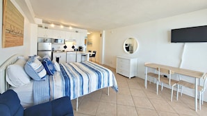 A compact bedroom with a bed, bedside table, and a small kitchenette. - Heated Oceanfront Pool + Hot Tub | Ocean View Studio | Free Daily Activities (Daytona Beach)