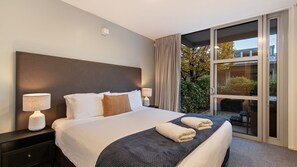 Ett modernt hotellrum med en stor säng, två sänglampor, en mörk sänggavel och en skjutdörr i glas som leder ut till en balkong med utsikt över träd och byggnader.