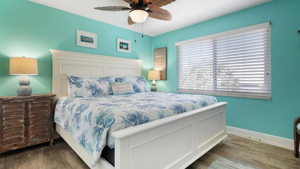 Une chambre à coucher avec un lit, un ventilateur de plafond, une fenêtre avec des stores et une table de chevet avec une lampe.