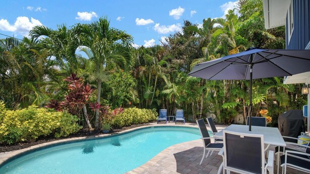 Une piscine dont le bord est revêtu de carrelage, entourée d’une végétation luxuriante et dotée d’un espace patio aménagé avec du mobilier d’extérieur.