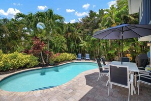 A swimming pool with a tiled edge, surrounded by lush greenery and a patio area with outdoor furniture. - Gone Coastal by Beach Boutique Rentals (Holmes Beach)