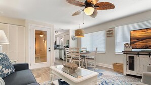 Un salon comprenant un canapé, une table basse avec un plateau en verre, un ventilateur de plafond et un téléviseur posé sur un meuble.