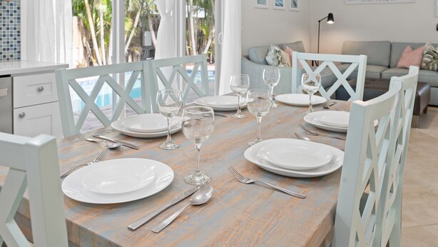 A dining table set with white plates, wine glasses, and cutlery, surrounded by white chairs with a blue and white pattern.