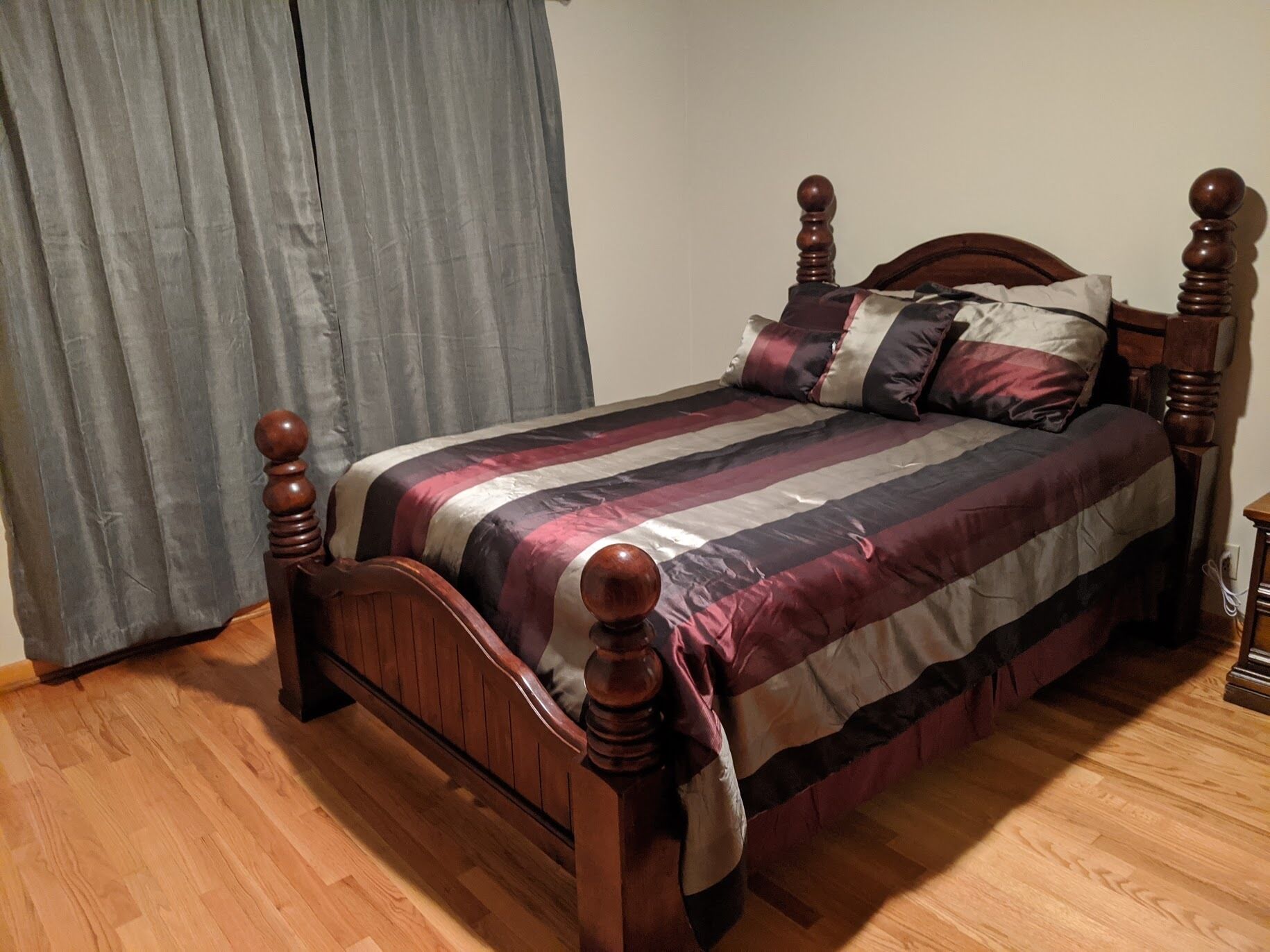 Un lit en bois avec une couvre-lit à rayures et deux oreillers, une table de chevet en bois et un rideau gris.