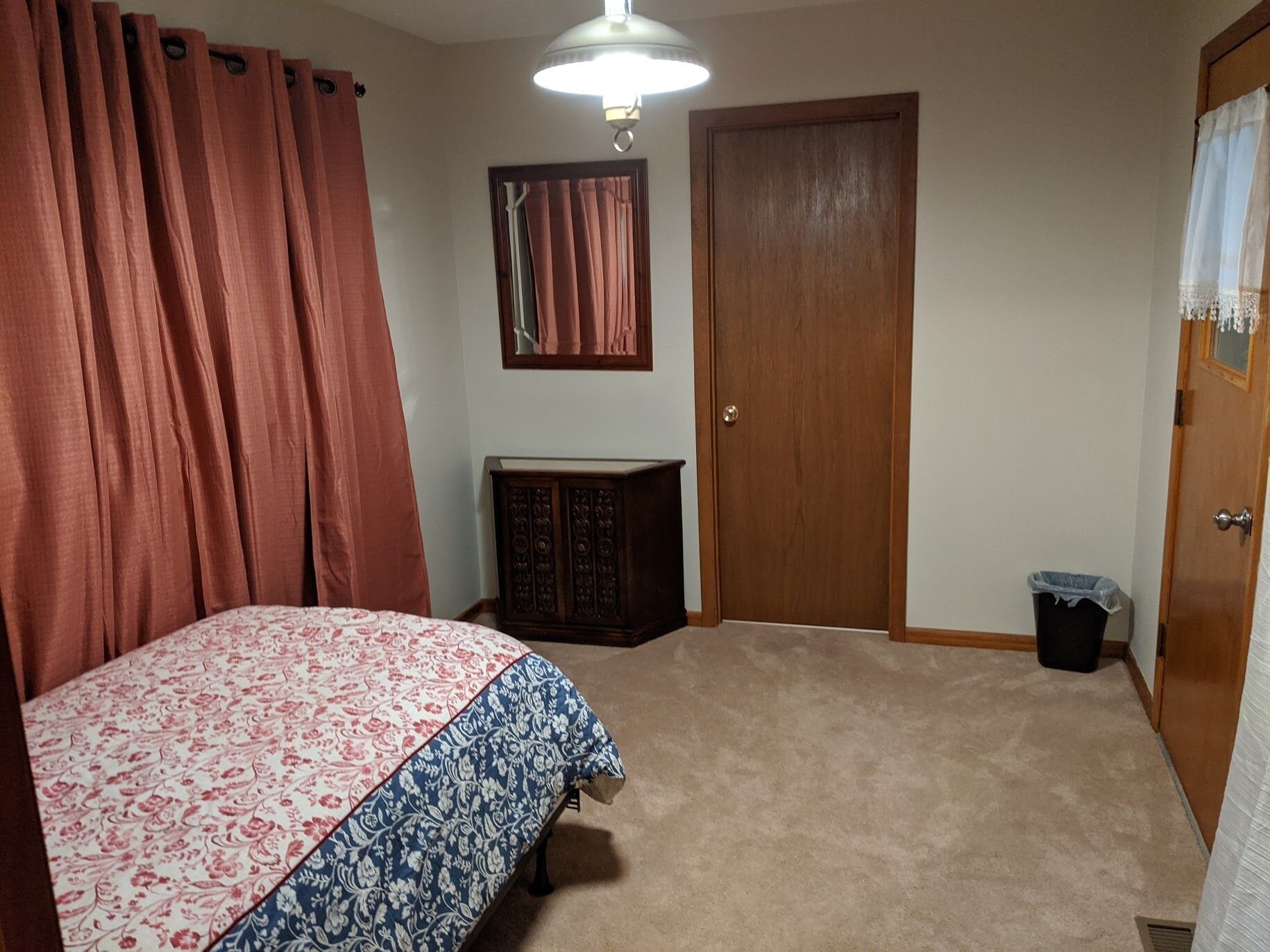 Ein Schlafzimmer mit einem Bett, einer Holztür, einer kleinen Kommode, einem Mülleimer und einer Deckenleuchte.