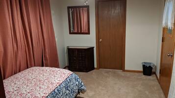 Ein Schlafzimmer mit einem Bett, einer Holztür, einer kleinen Kommode, einem Mülleimer und einer Deckenleuchte.