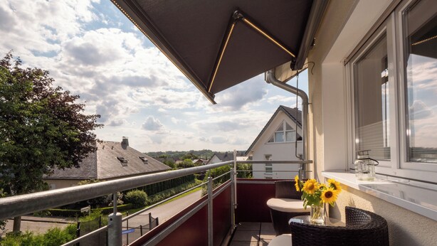 En balkon med udsigt til huse, en fletstol, et lille bord og en vase med blomster.