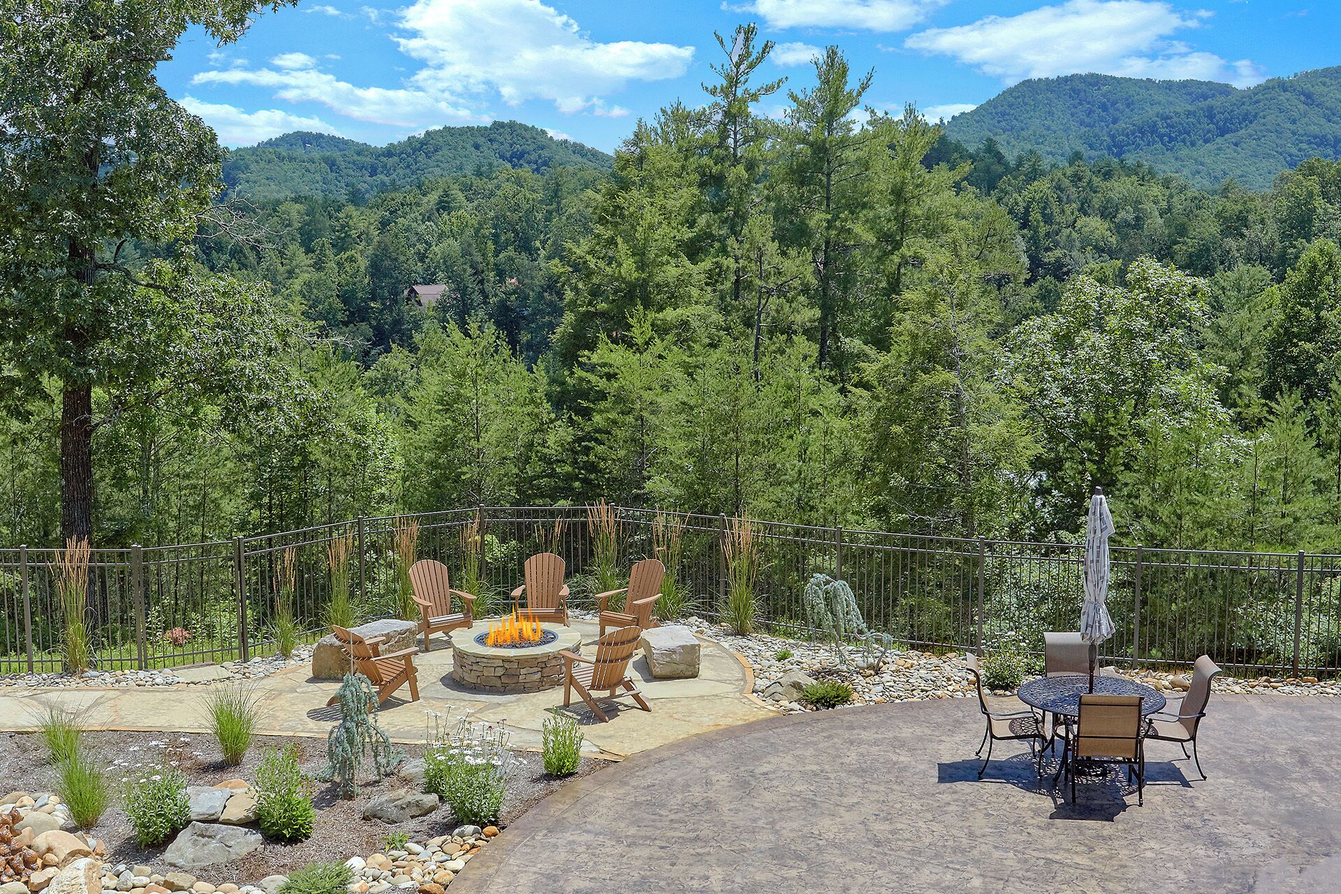 An outdoor patio with a stone fire pit, wooden chairs, and a table with an umbrella.