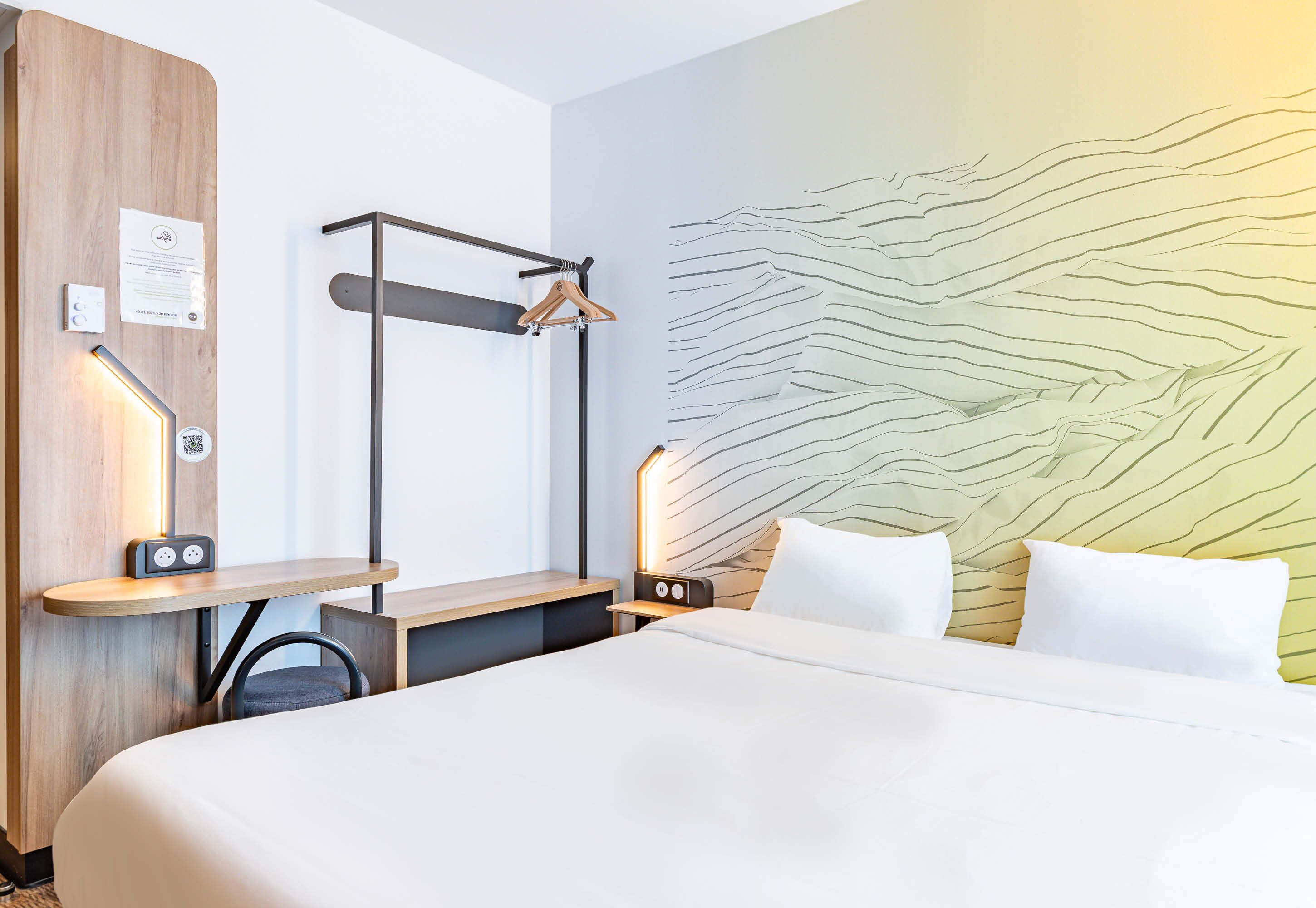 Habitación de hotel con cama, mesitas de noche, escritorio, silla, armario, pared con diseño texturizado y perchero.