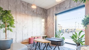 A modern interior with a concrete wall, a large window, a seating area with a table, and potted plants. - B&B HOTEL Poitiers Aéroport (Biard)