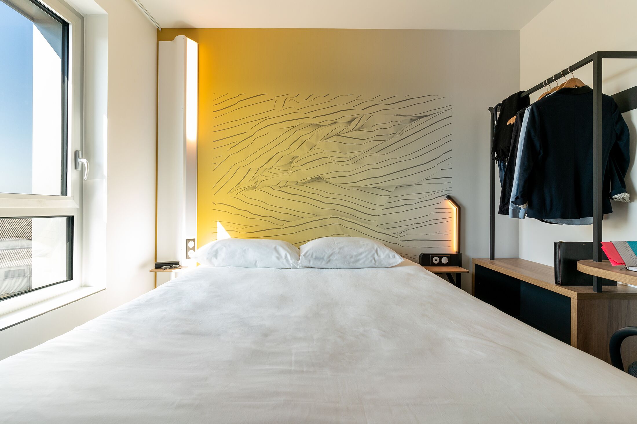 A hotel room with a bed, a bedside table, a lamp, a wardrobe, and a wall with a unique design.
