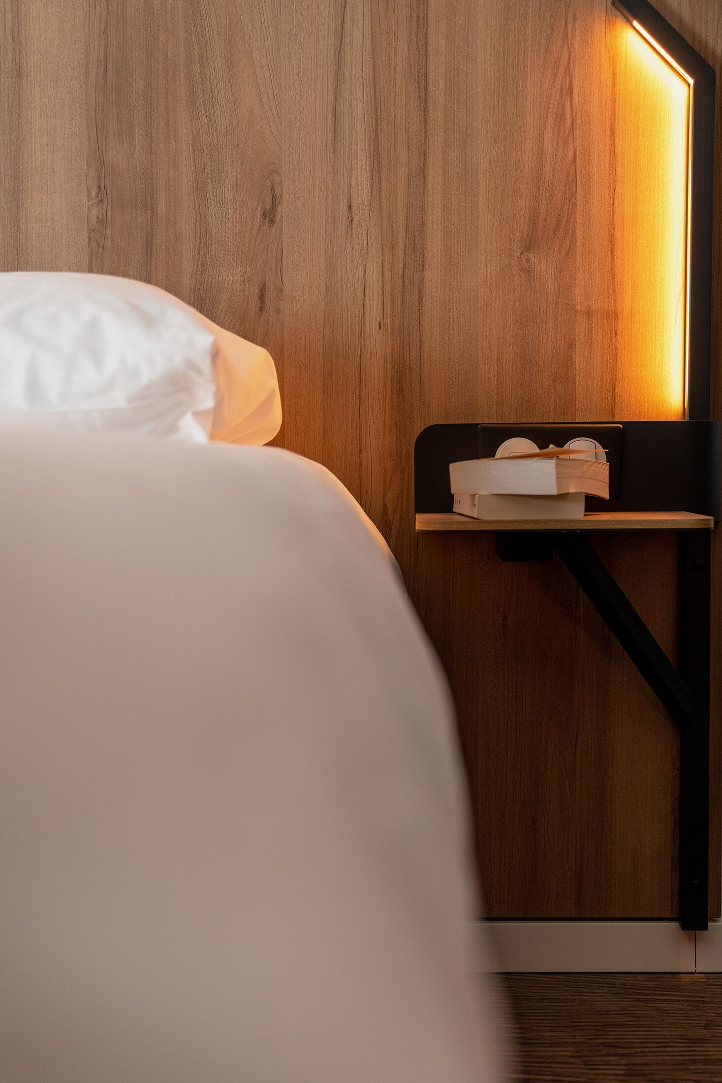 A neatly made bed with white linens, a bedside table with a book, and a wall-mounted light fixture.