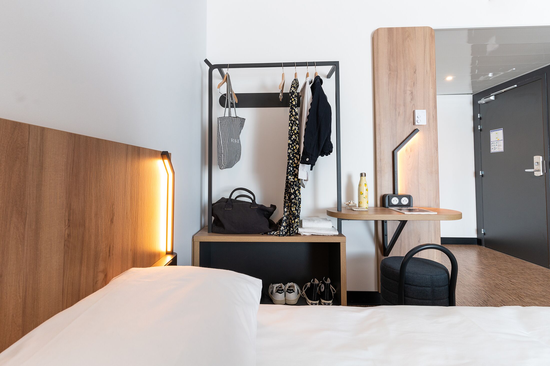 A hotel room with a bed, a wooden headboard, a small table with a lamp, a chair, a coat rack with clothes and a bag, and a shoe rack with shoes.