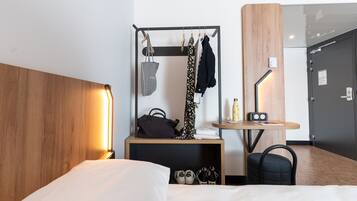 A hotel room with a bed, a wooden headboard, a small table with a lamp, a chair, a coat rack with clothes and a bag, and a shoe rack with shoes.