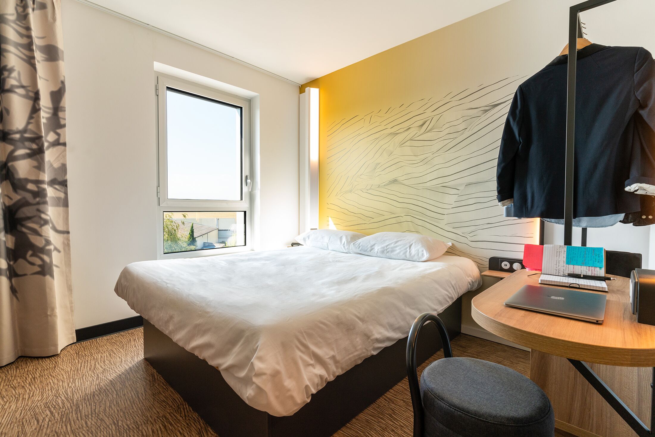 A hotel room with a bed, a desk with a laptop, a chair, a window, and a curtain.