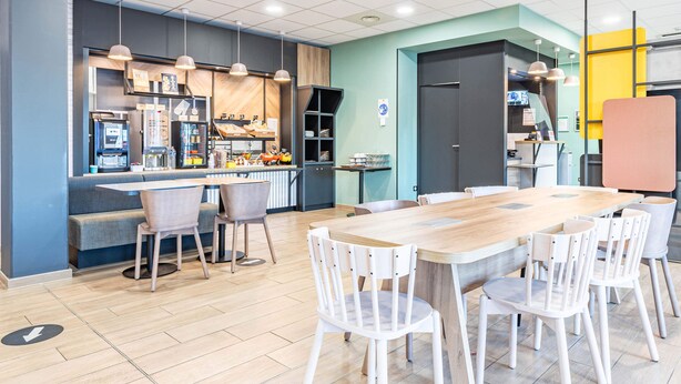 A modern dining area with a long wooden table and white chairs, a counter with a coffee machine, and a seating area with a bench. - B&B HOTEL Poitiers Aéroport (Biard)