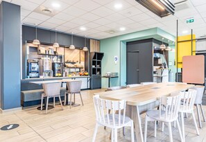 A modern dining area with a long wooden table and white chairs, a counter with a coffee machine, and a seating area with a bench. - B&B HOTEL Poitiers Aéroport (Biard)