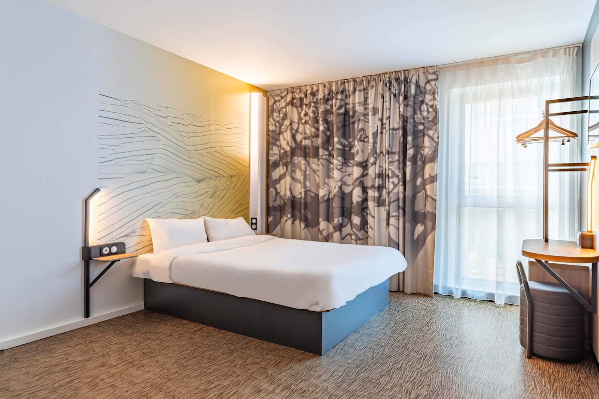 A modern hotel room with a large bed, a bedside table with a lamp, a wardrobe, and a view of the outdoors through sheer curtains.