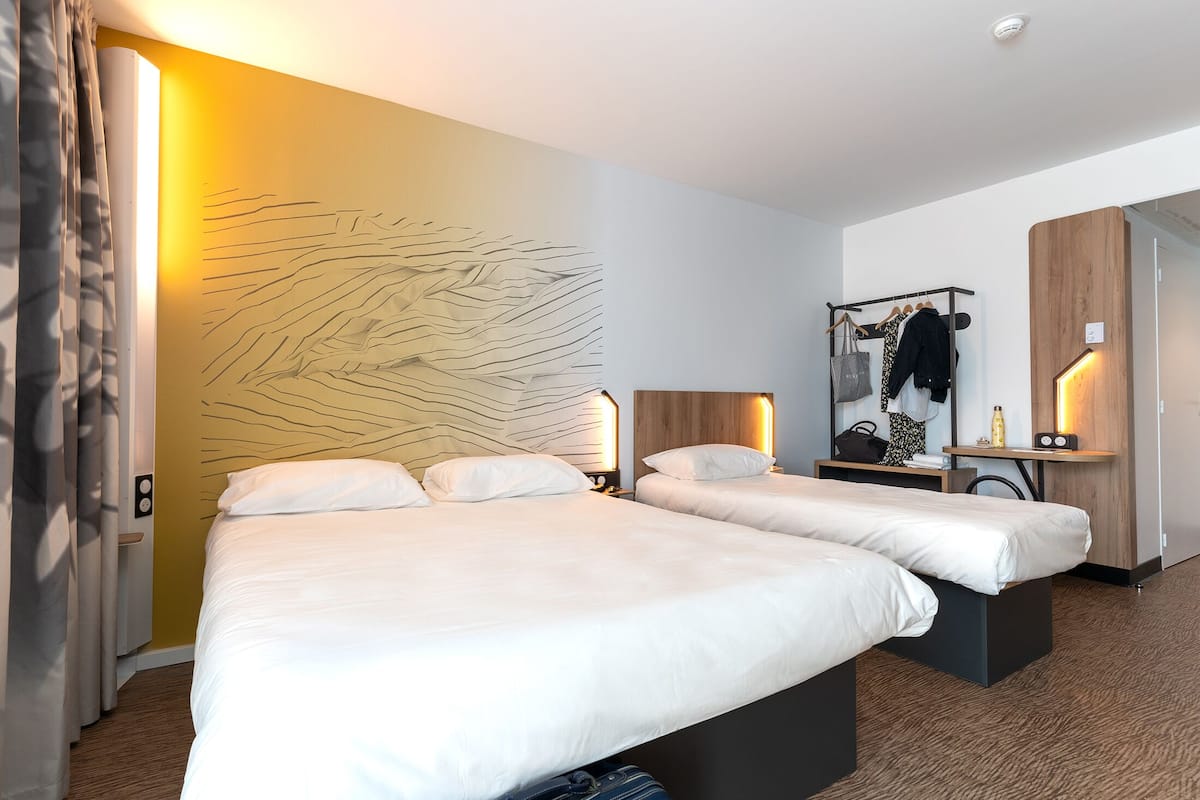 A hotel room with two beds, a wooden headboard, a wall art piece, a bedside table with a lamp, and a coat rack with clothes and a bag.