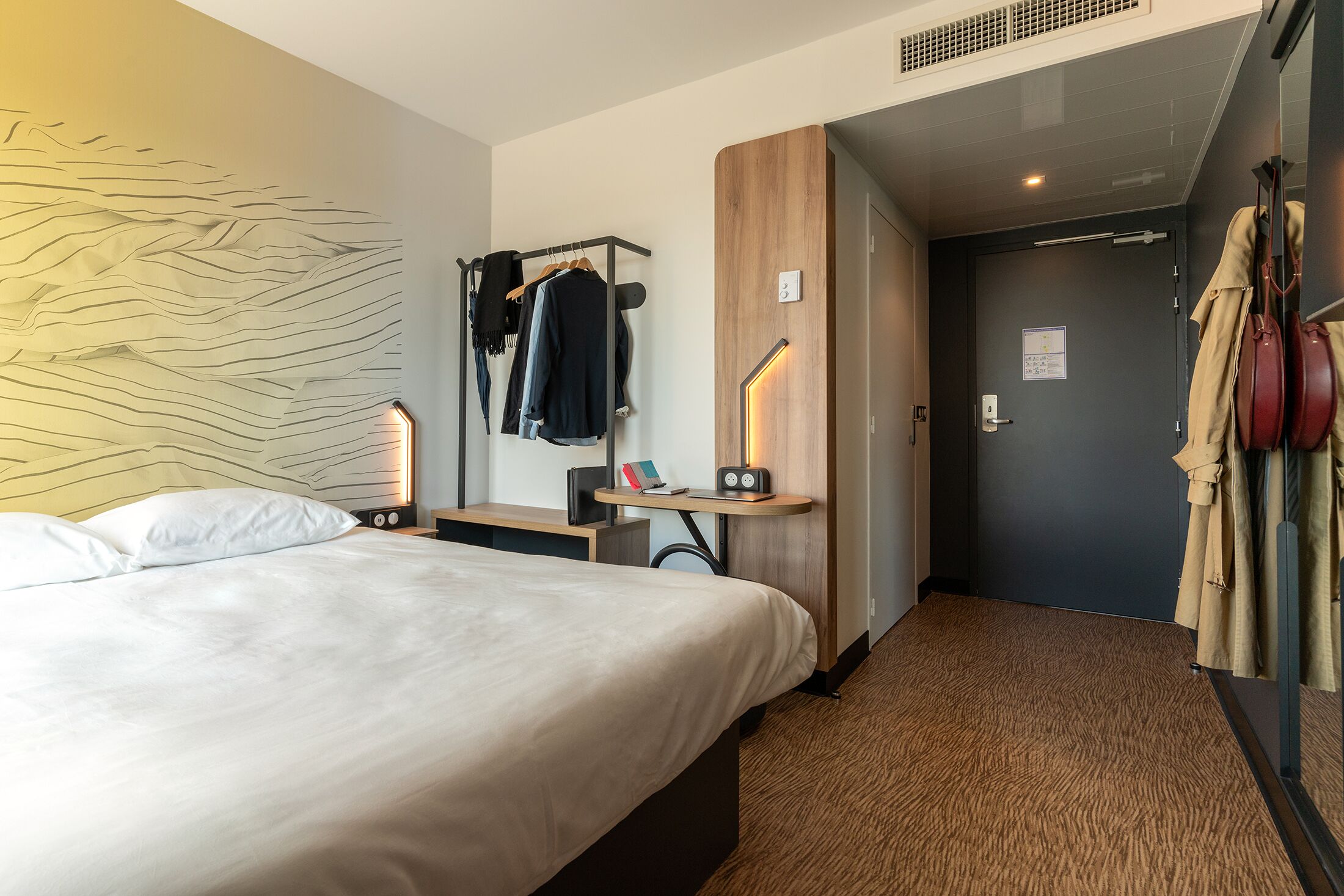 A hotel room with a bed, a desk with a lamp, a coat rack, and a door leading to another room.