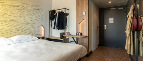 Quarto de hotel com uma cama, uma escrivaninha com candeeiro, um cabideiro e uma porta que dá para outro quarto.