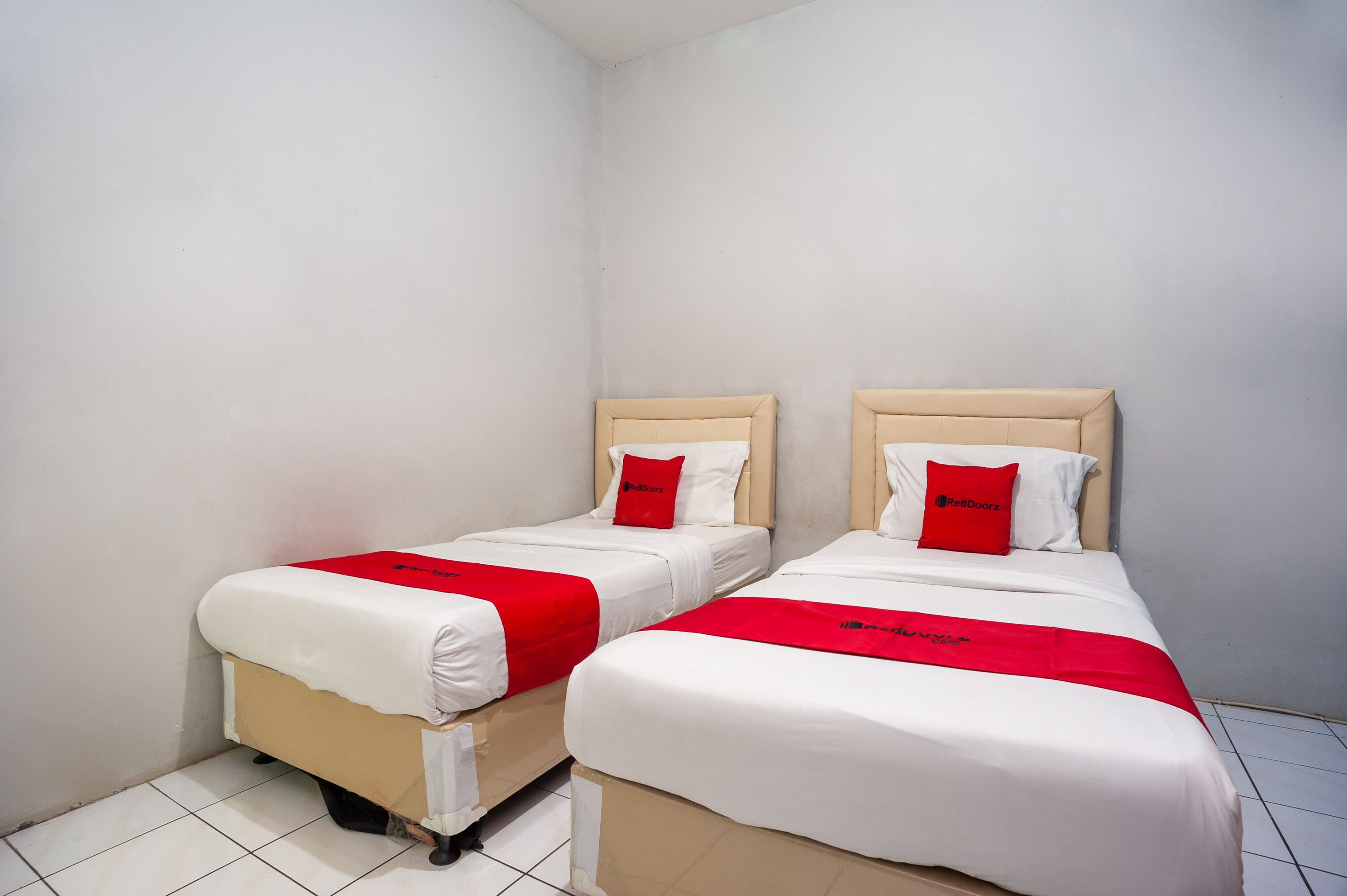 Two single beds with white bedding and red accents, in a corner room with white walls and tiled flooring.