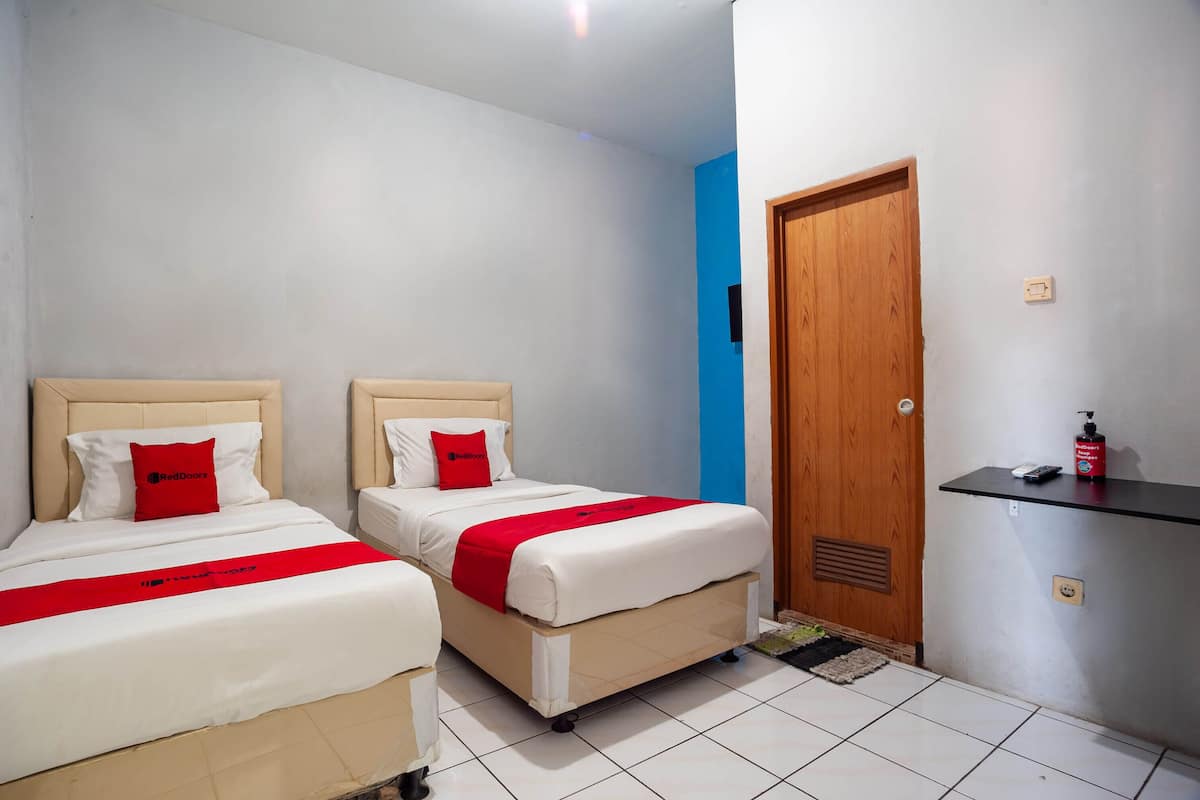 Une chambre d’hôtel avec deux lits, chacun agrémenté d’un oreiller rouge et d’une couverture blanche. On y trouve une porte en bois, une petite étagère avec une bouteille, et un mur d’accent bleu.
