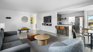 A modern living room with a sofa, a coffee table, and a TV. There is a kitchen area with a refrigerator, a sink, and a bar. The room has a large window and a view of the outdoors.