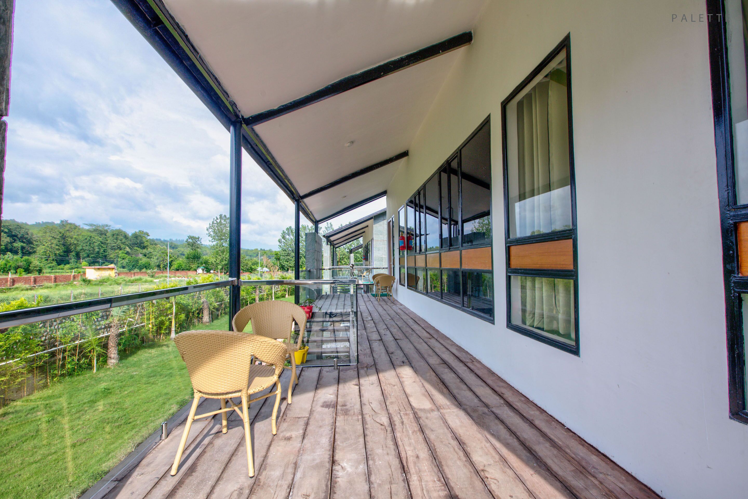 A covered balcony with wooden flooring, wicker chairs, and a glass railing overlooking a grassy area.