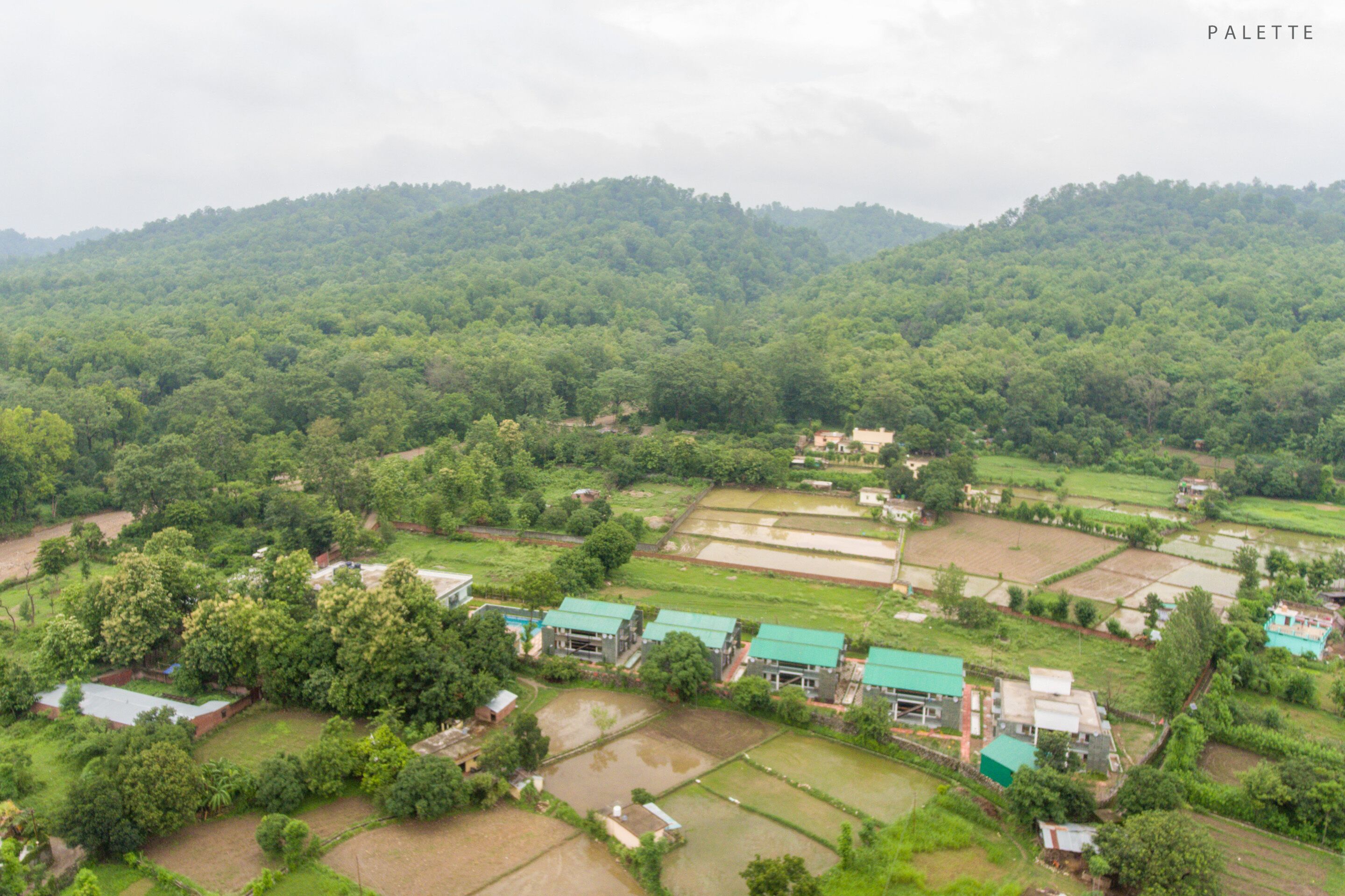 An aerial view of a rural village with green fields, scattered houses, and a hilly backdrop.