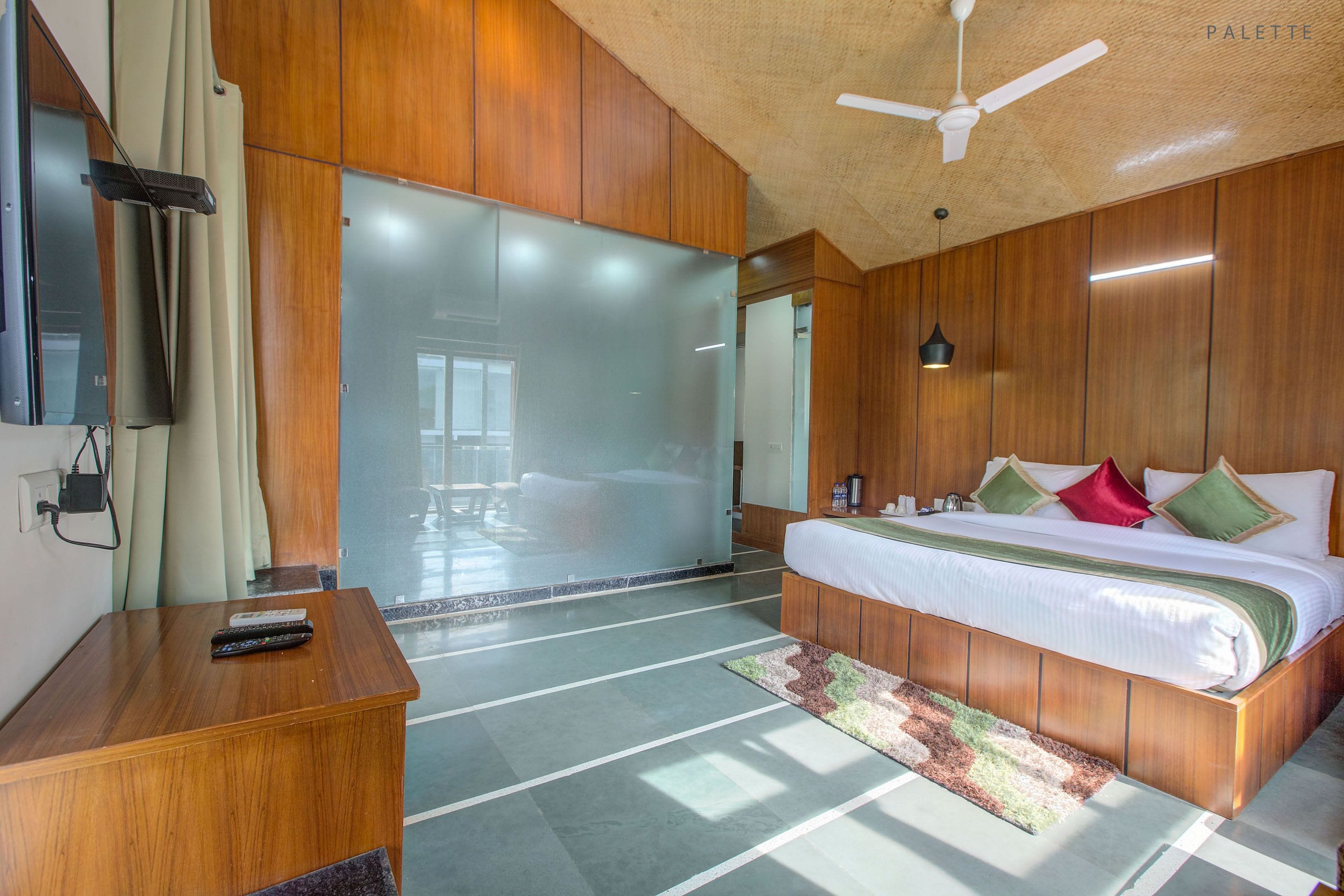 Habitació d'hotel amb sòl de fusta, un llit amb almoades, escriptori amb comandament a distància i ventilador de sostre.