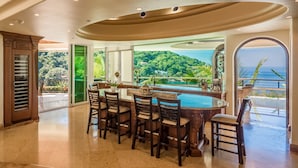 A spacious kitchen with a large island, a dining area with chairs, and a view of a lush landscape and ocean. - Fully Staffed, Beach frontage Luxury Villa (Boca de Tomatlán)