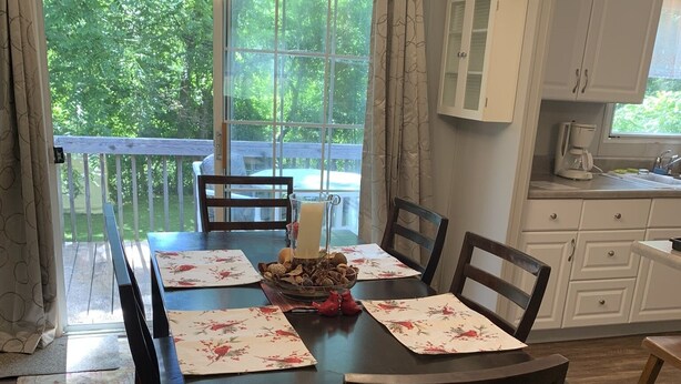 A dining room with a large table set for six, surrounded by dark wooden chairs. A chandelier hangs above, and a window offers a view of greenery outside. - Cardinal  Cottage 55 Patton Cemetery Road (Swannanoa)