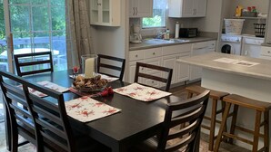 A dining area with a dark wooden table, matching chairs, and a chandelier. A kitchen is visible in the background with white cabinets and a window. - Cardinal  Cottage 55 Patton Cemetery Road (Swannanoa)