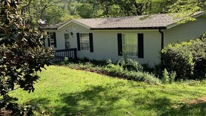 A single-story house with a sloped roof, surrounded by a well-maintained lawn and dense trees. - Cardinal  Cottage 55 Patton Cemetery Road (Swannanoa)