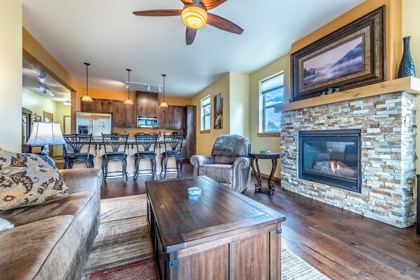 A spacious living room with a fireplace, a dining area with a kitchen in the background, and a ceiling fan.