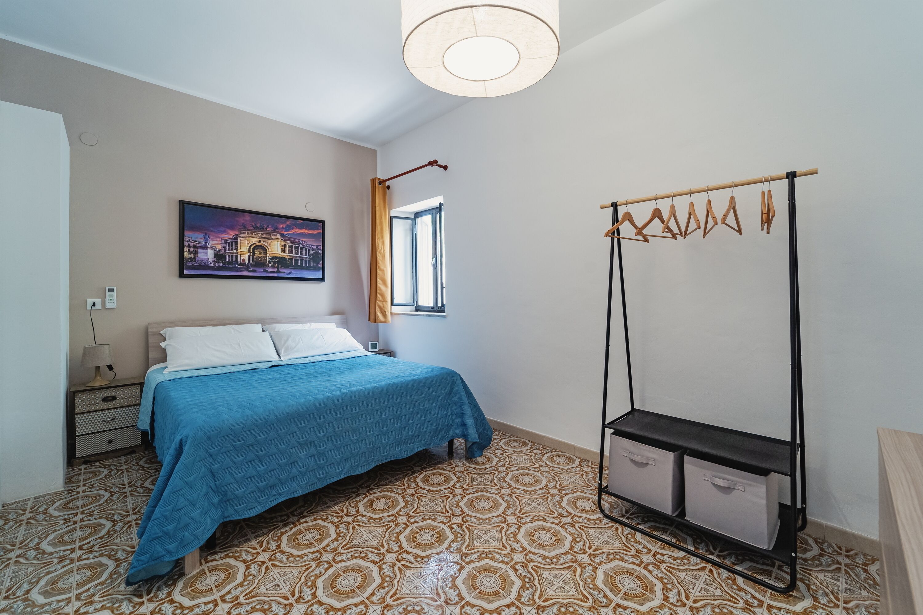 A bedroom with a bed, a painting, a window, a wardrobe, and patterned flooring.