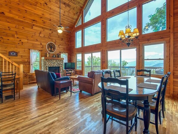 A spacious wooden cabin interior with a dining area, fireplace, and large windows offering scenic views. - 5 BD 3 BA Log Cabin Sleep 13 Long Range Views Hot Tub, Game Room Wifi Garage (Waynesville)