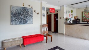 A hotel lobby with a reception desk, a bench with a red cushion, a large abstract painting, and a chandelier.